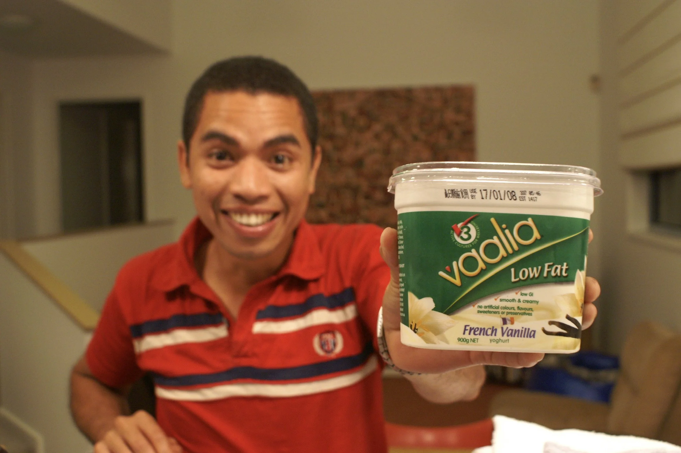 A smiling man in a red shirt holding a container of Vadalia low-fat French vanilla yogurt towards the camera in a living room.