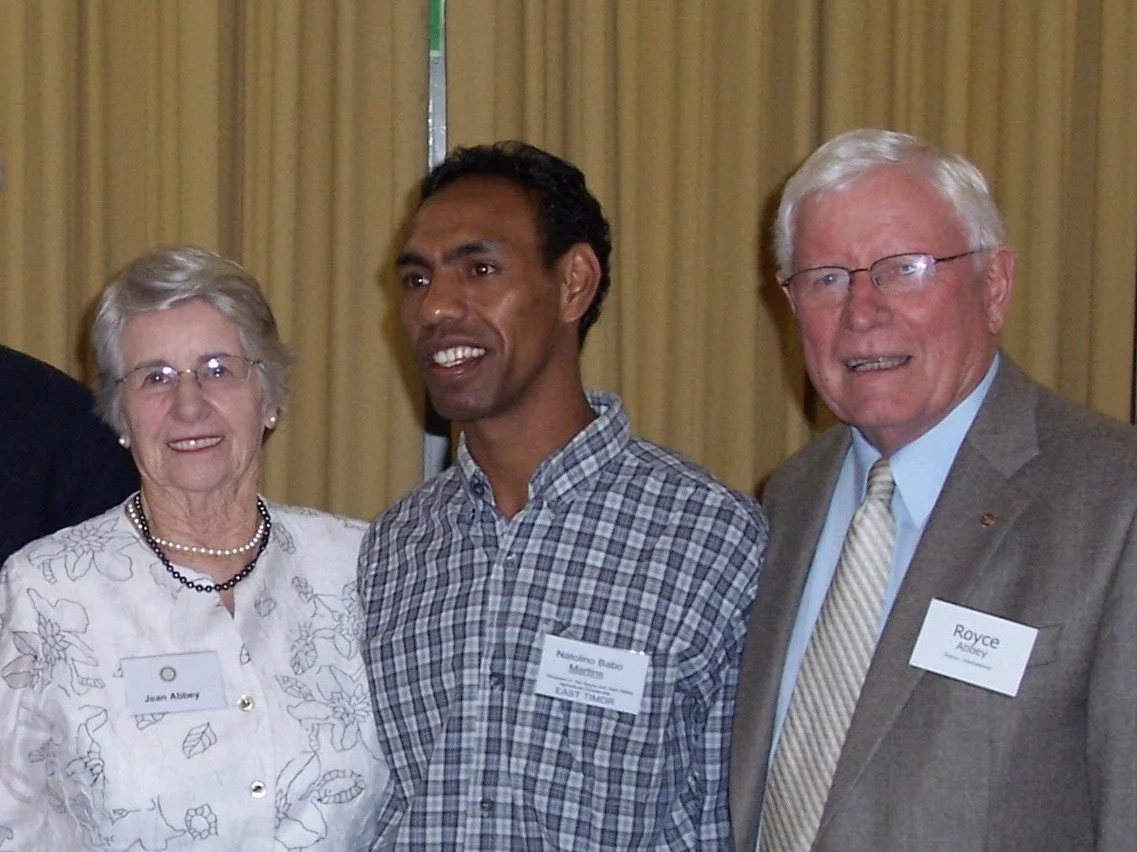 Three people standing together at an indoor event, smiling for the camera. The woman on the left is wearing glasses and a white floral blouse with a pearl necklace. The man on the right has white hair, glasses, and is dressed in a gray suit with a li
