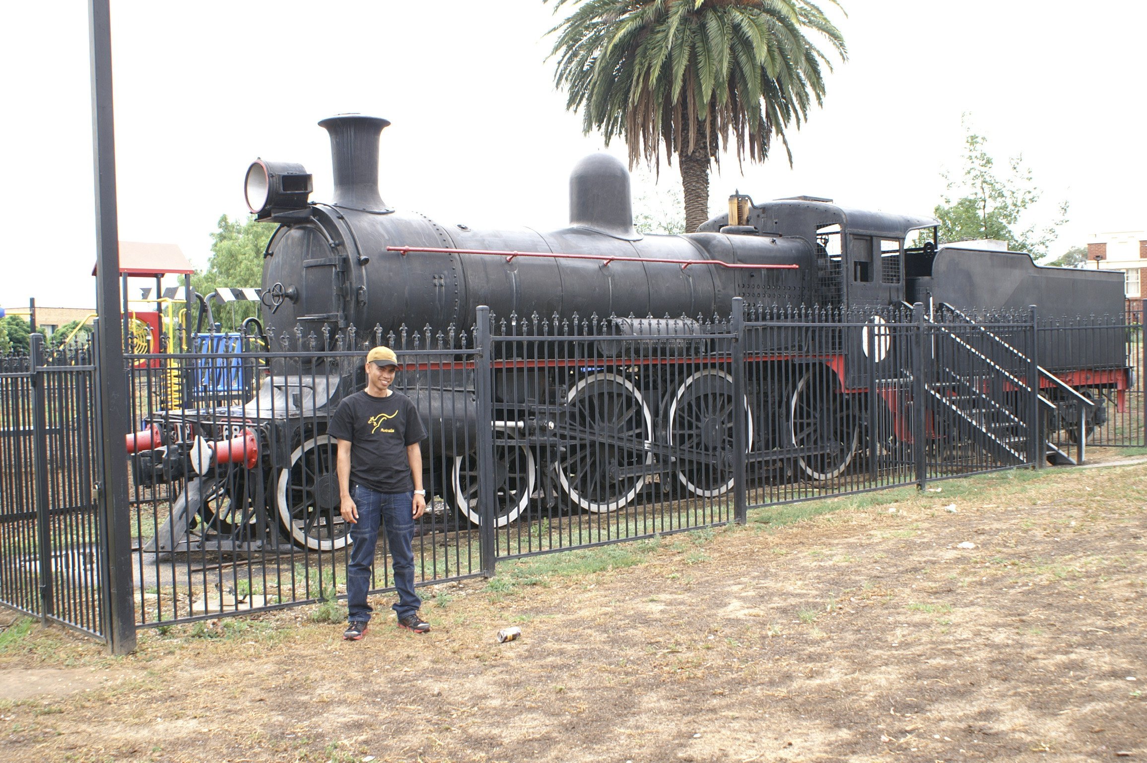 A smiling man wearing a black T-shirts, blue jeans, black sneakers, and a yellow cap stands in front of a vintage black steam locomotive enclosed by a black metal fence in a park. A large palm tree is visible behind the train.