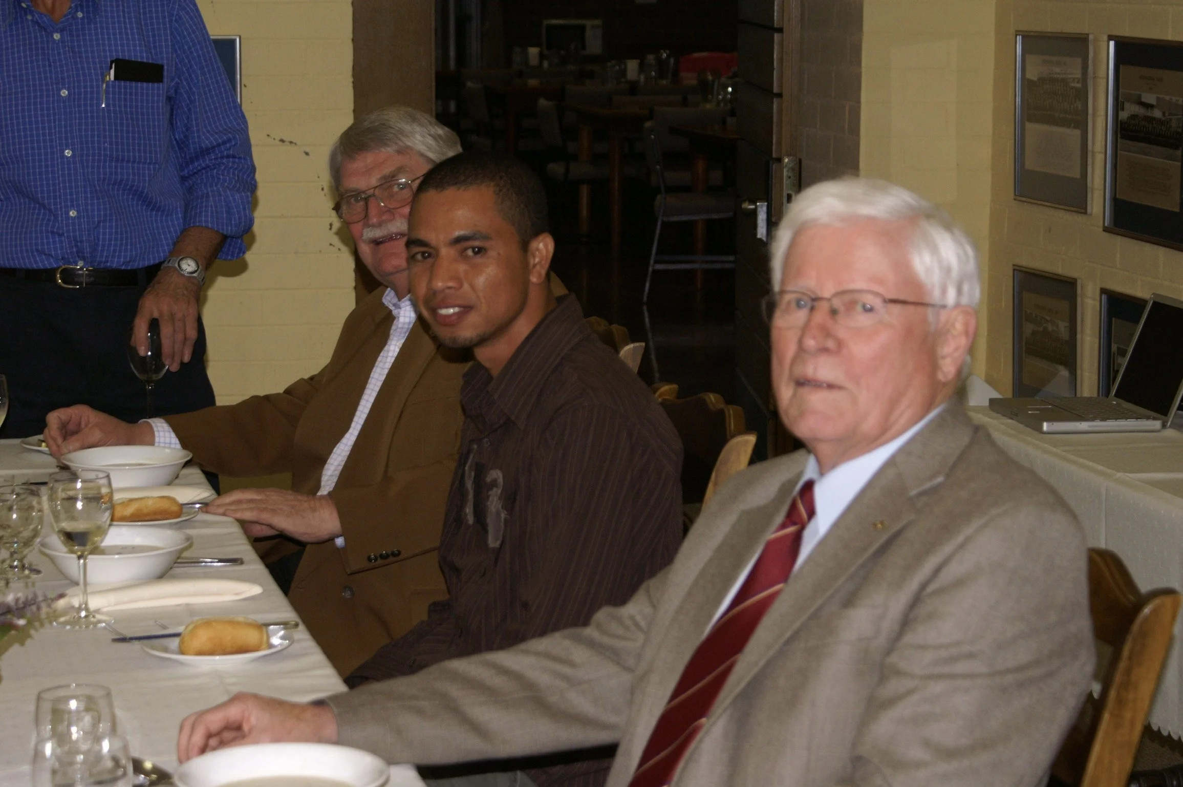 Four men sitting at a dining table with plates, glasses, and bread rolls, in a restaurant or banquet setting.