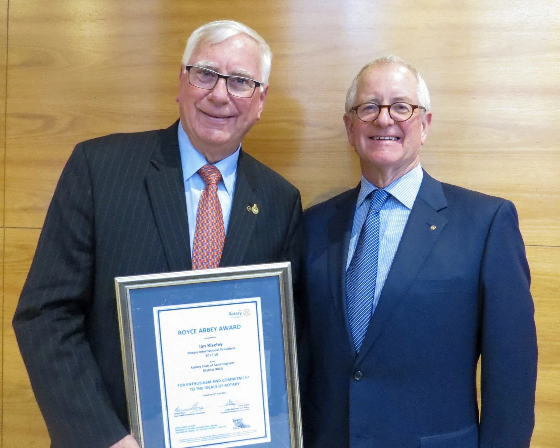 Two men in suits smiling, one man holding a framed award standing in front of a wooden wall.