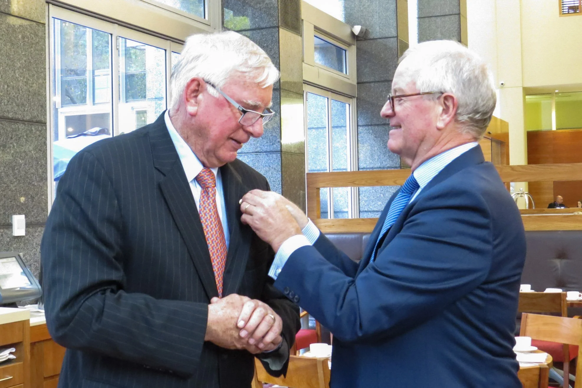 Two elderly men in suits, one pinning a medal on the other, inside a modern building with large windows and wooden paneling.
