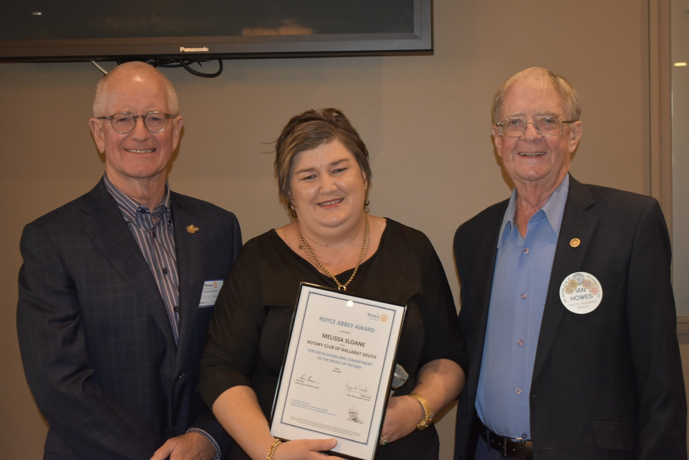 Three people standing together, with the woman in the middle holding a certificate. All are smiling, wearing business attire, and have name tags or pins.