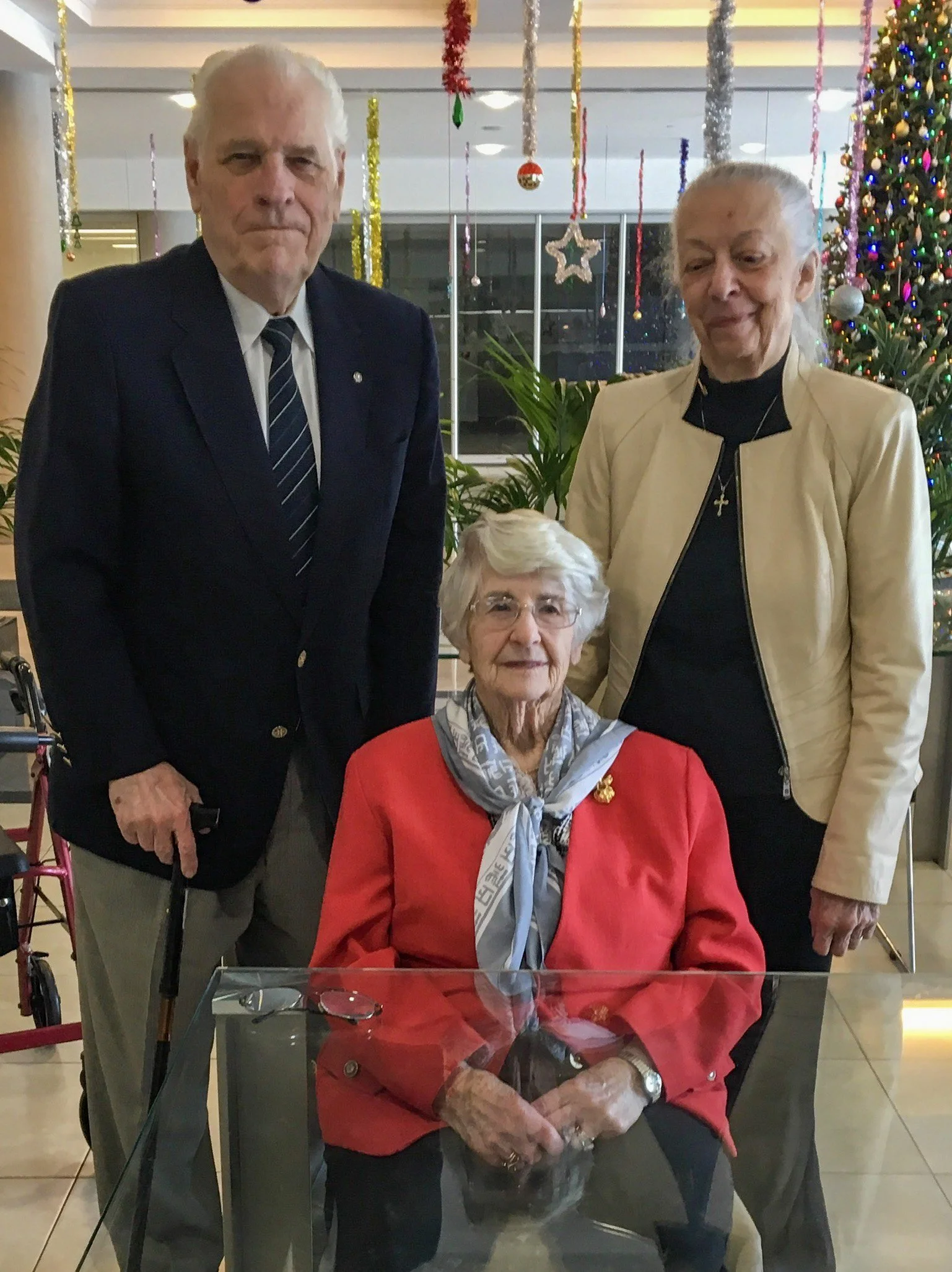 omen and one elderly man posing for a photo in front of Christmas decorations, including a decorated Christmas tree and hanging ornaments. One woman is seated in a wheelchair, wearing a red blazer and scarf, while the other two women and the man stan