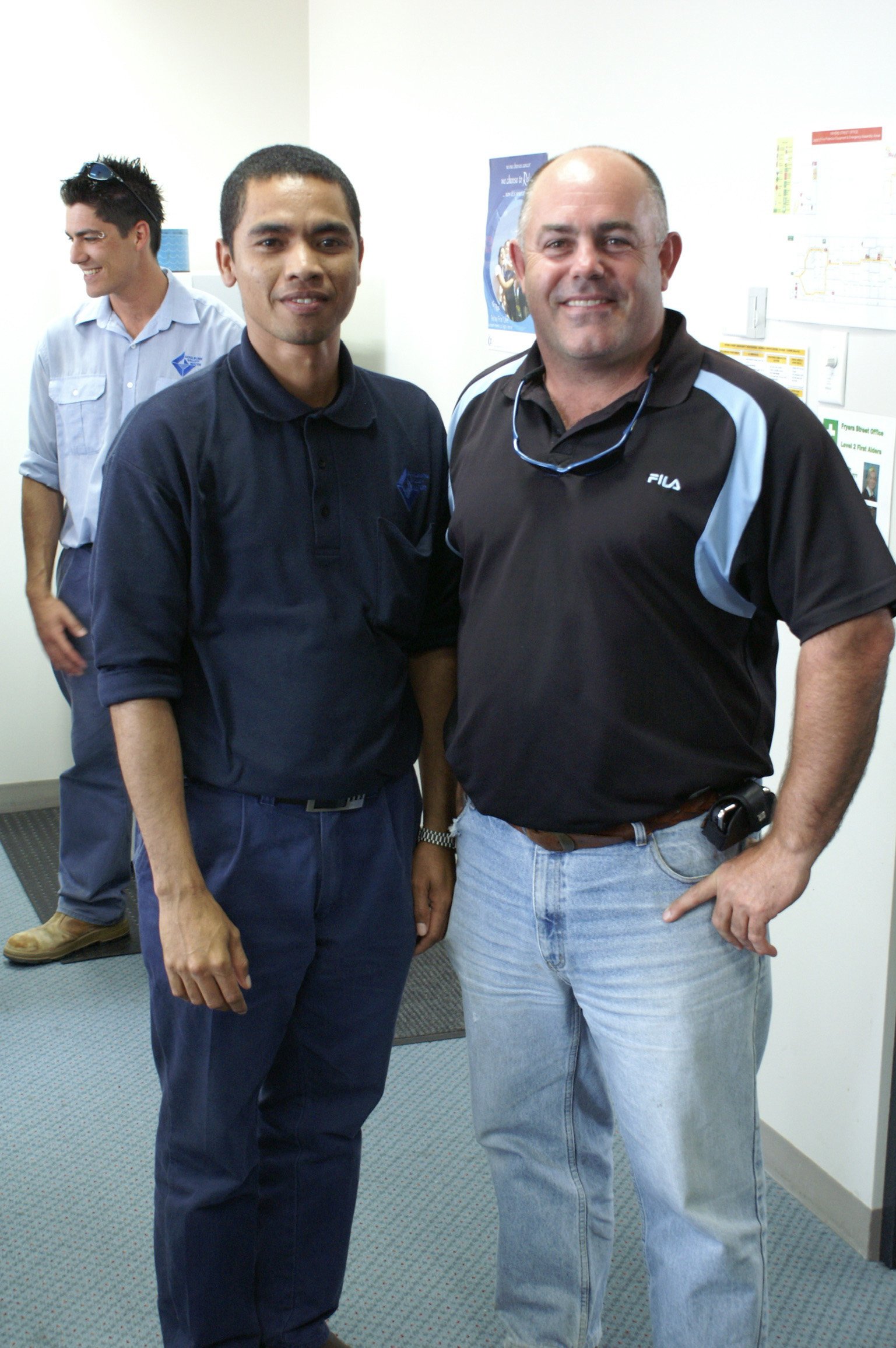 Two men standing close together smiling at the camera in an indoor setting, with a third man standing in the background. All are casually dressed, and there are posters and informational signs on the wall behind them.