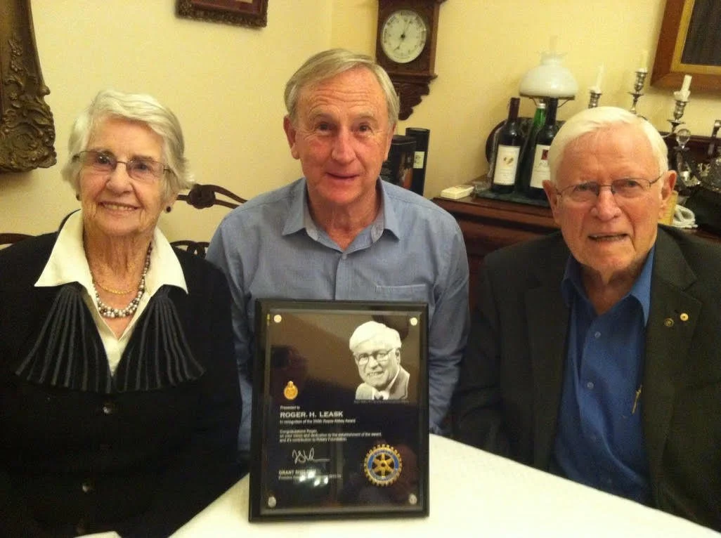 Three elderly individuals sit at a table, posing with a framed award for Roger H. Leak, with a woman on the left, a man in the middle, and another man on the right. The background shows a clock, bottles of wine, and decorative items.