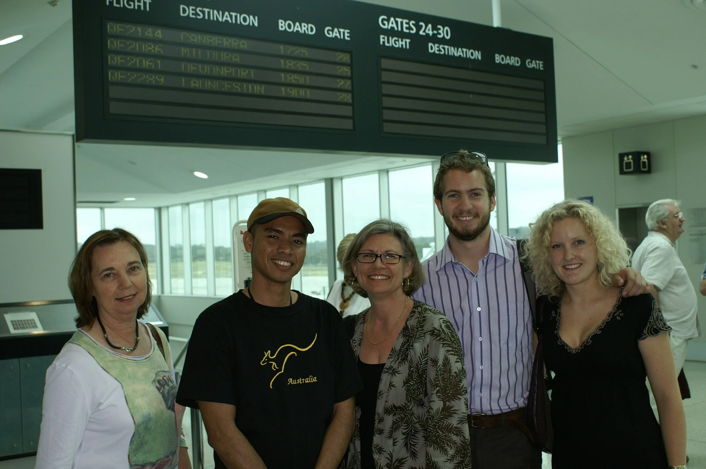 A group of five smiling people standing together in an airport terminal, with flight information board above them.
