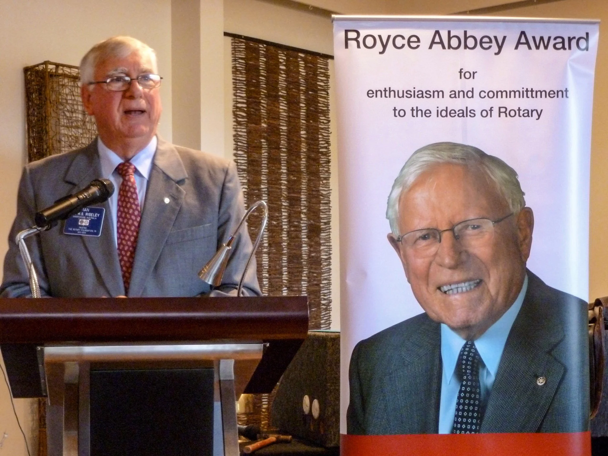 An elderly man with glasses and gray hair, wearing a gray suit and a red patterned tie, standing at a podium with a microphone, speaking. Next to him is a large vertical banner with a picture of another elderly man with glasses and gray hair, wearing