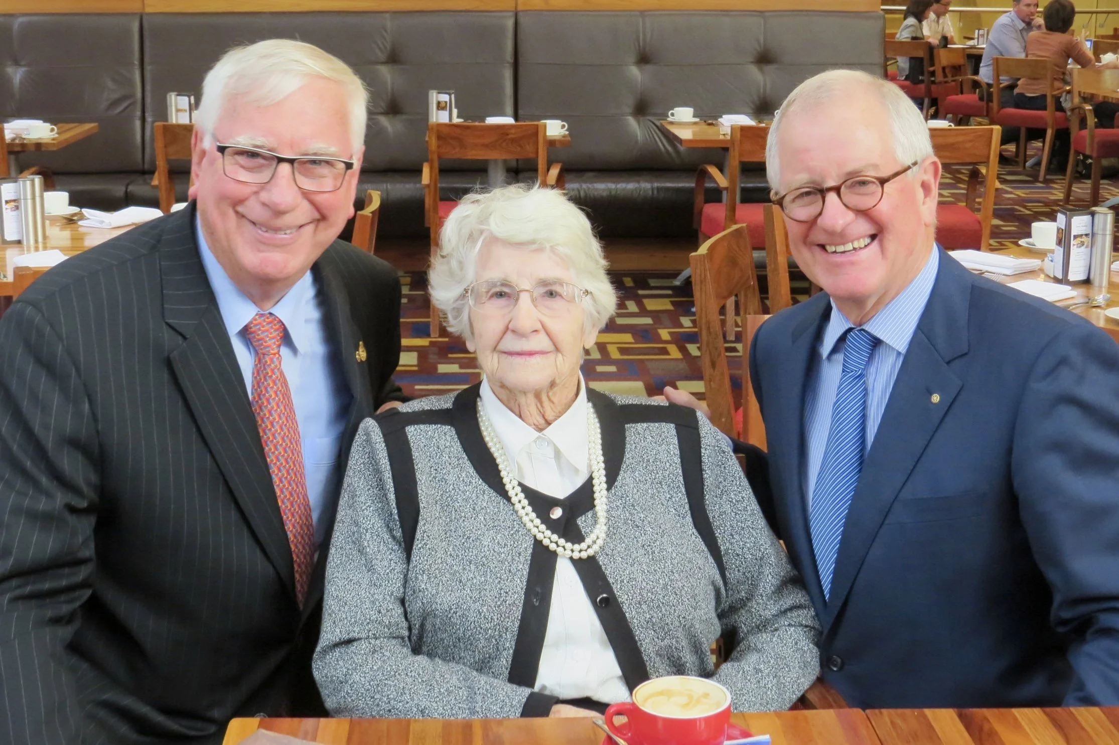 Three smiling elderly individuals, two men in suits and a woman in a sweater, are gathered around a table in a restaurant or cafe. The woman has white hair and is wearing glasses, a white blouse, a necklace, and a gray cardigan. The men have white ha