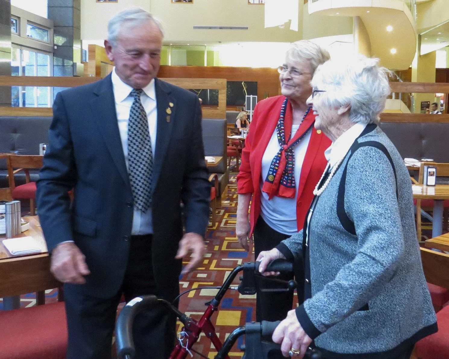 Three elderly women and a man standing in a restaurant, with two women talking to the man. One woman is using a walker.