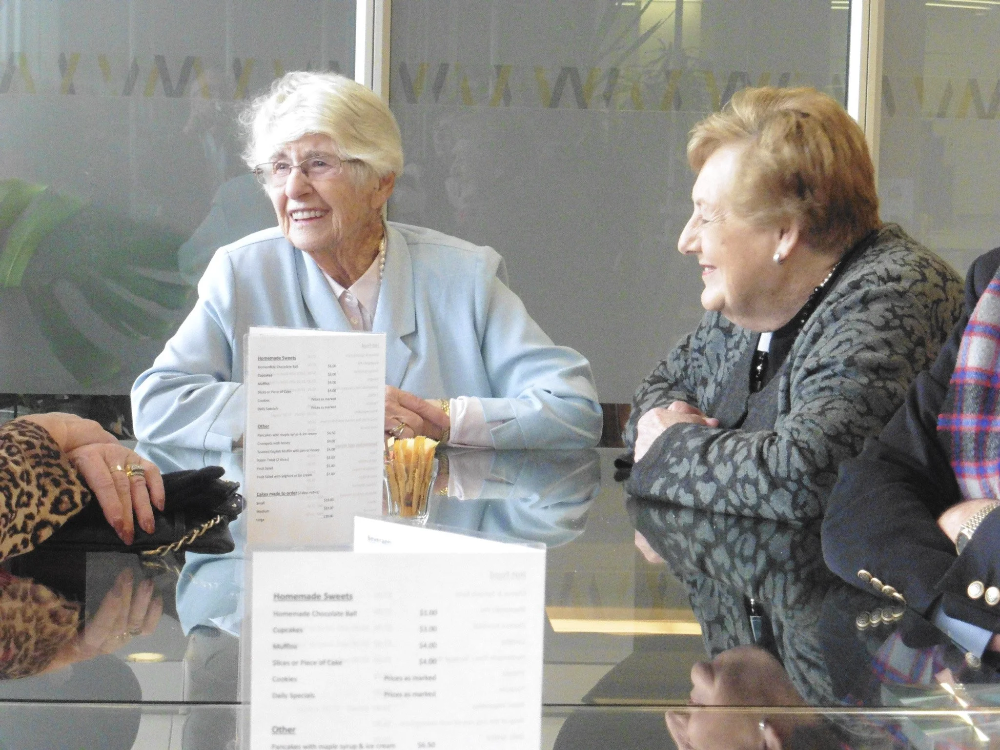 Four elderly women sitting around a glass table, smiling and engaging in conversation in a bright indoor setting, with menus and a container of straw sticks on the table.