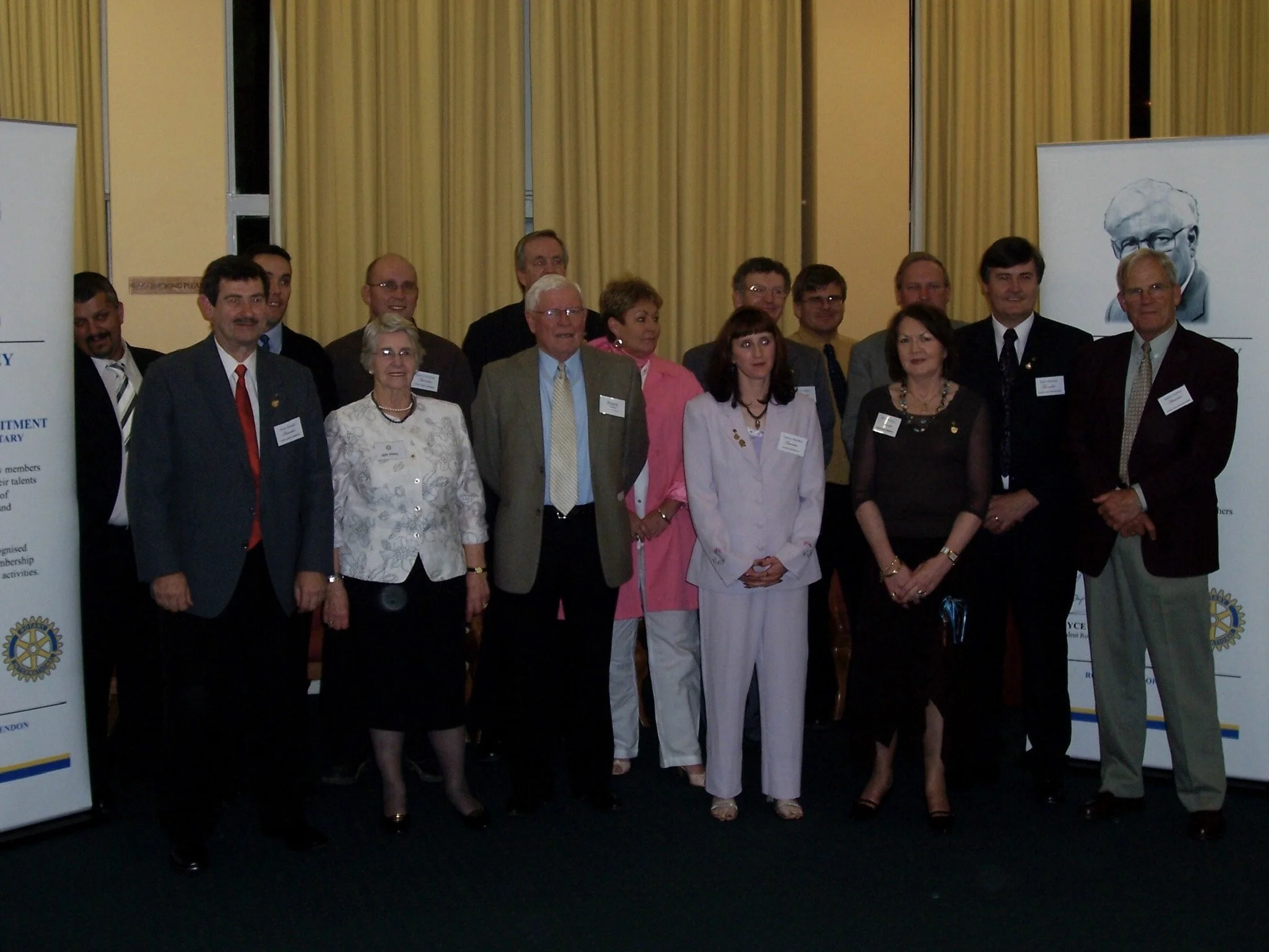 Group of people standing together at an indoor event, some wearing name tags, with two banners displaying Rotary International logos on either side.