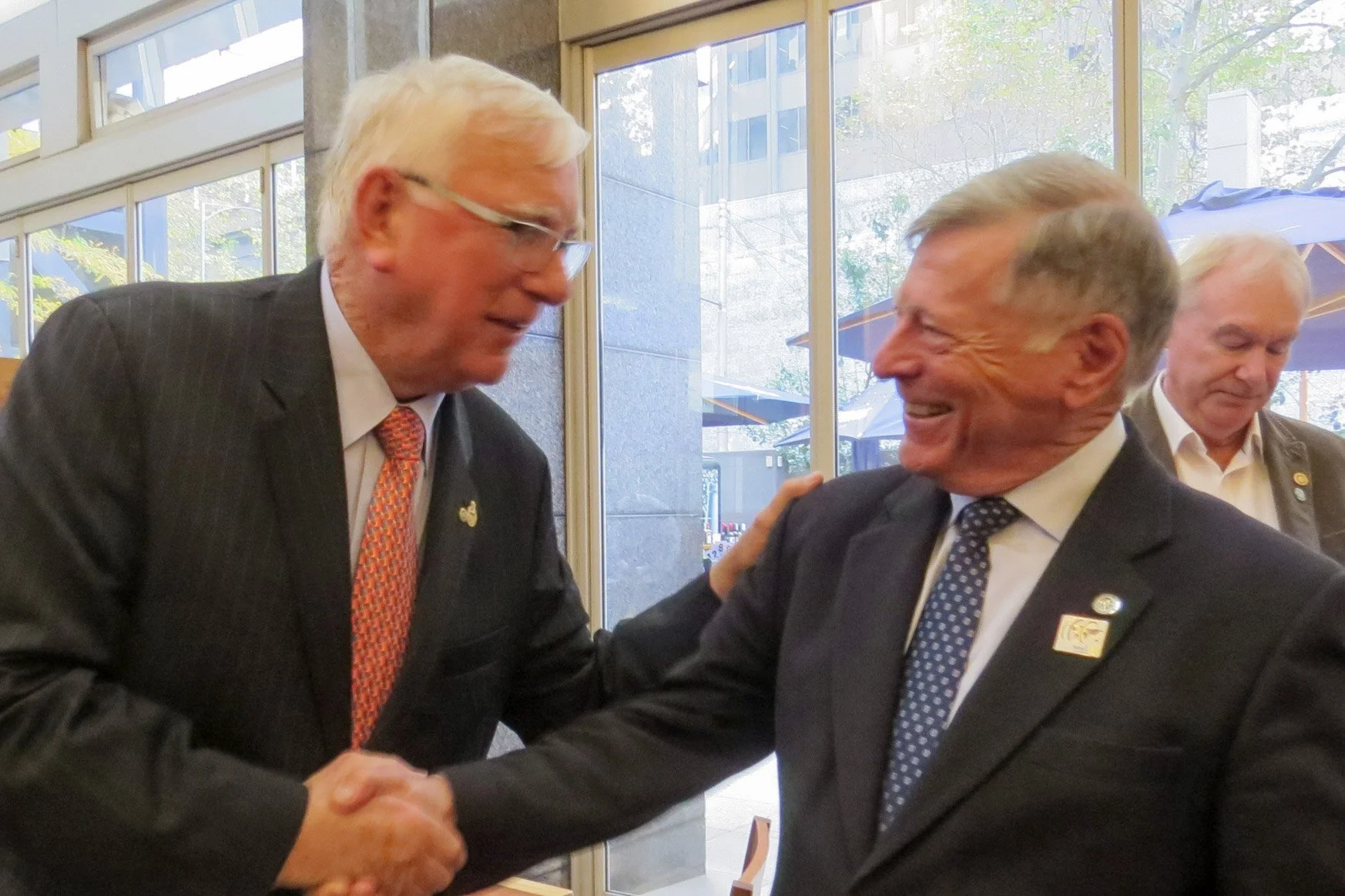 Two men in suits shaking hands and smiling in a professional setting with windows and other people in the background.