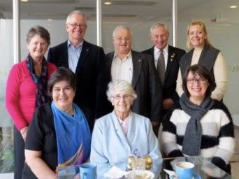 Group of seven people, five women and two men, gathered together in an indoor setting, smiling for the photo. One woman in a wheelchair is seated in front.