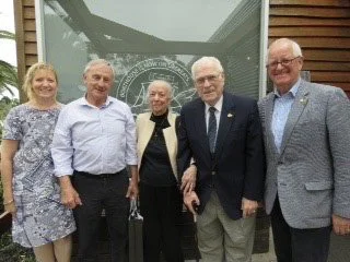 Five adults standing outside in front of a window and building. Two women and three men, all smiling and dressed in semi-formal attire.