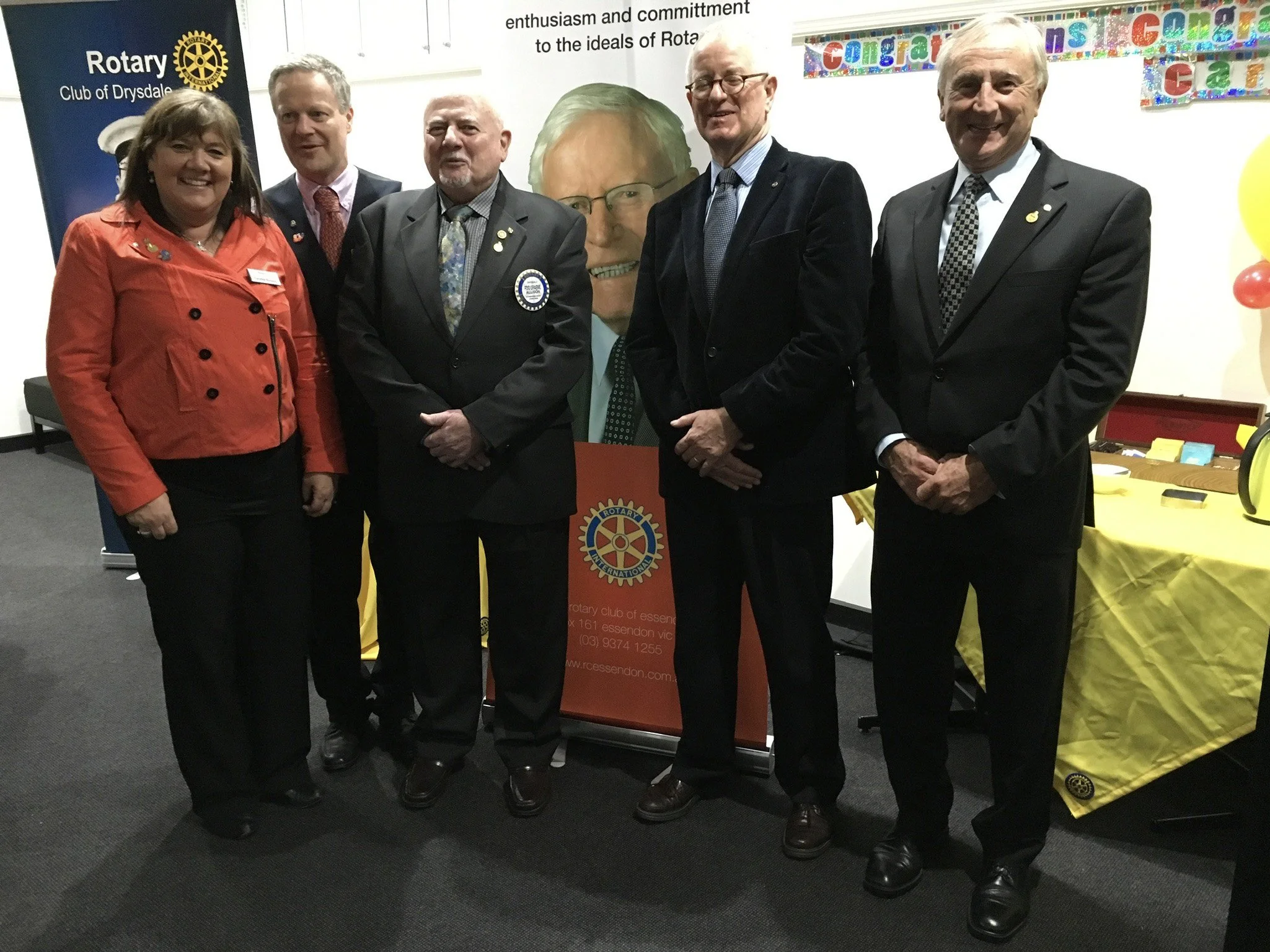 Group of six people standing together at a Rotary Club event, with banners and colorful decorations in the background.
