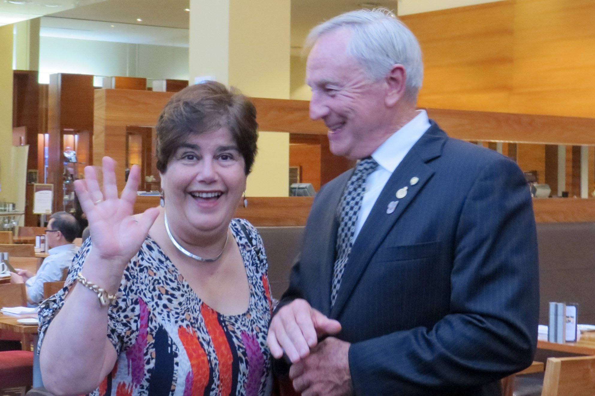A woman with short brown hair and earrings, smiling and waving, standing next to a man with gray hair, smiling and looking at her, in a restaurant or cafe setting.