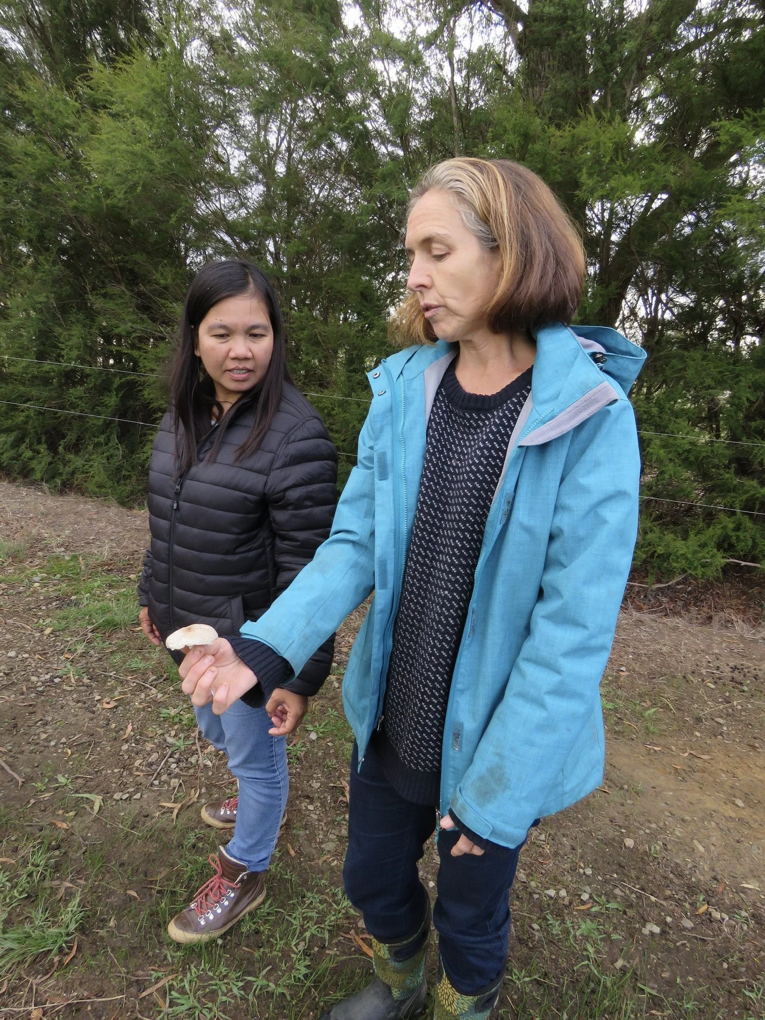 Two women outdoors, one holding a small mushroom and the other looking at it, with trees and an overcast sky in the background.