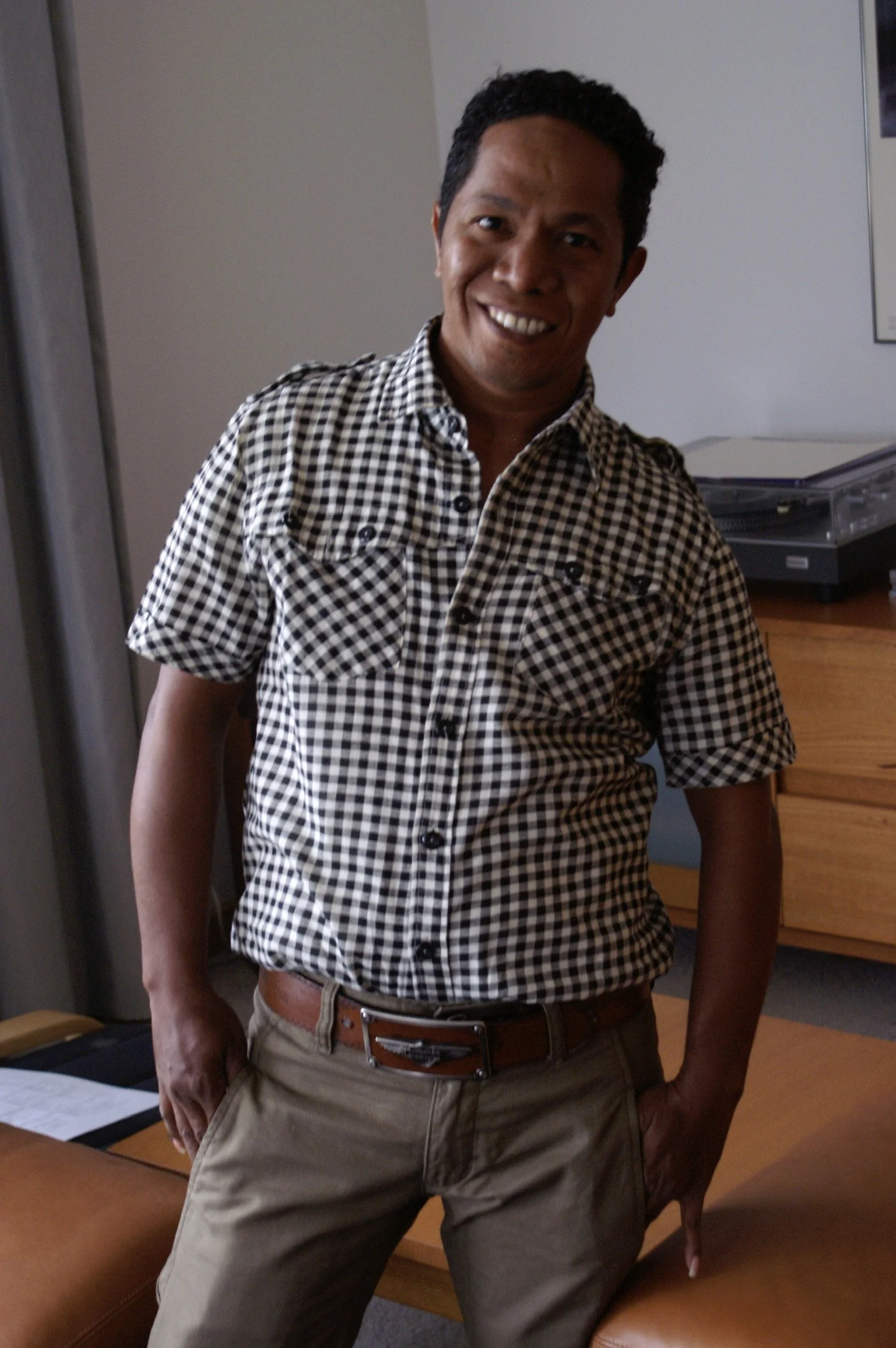 A man with curly black hair wearing a black and white checkered shirt and khaki pants standing in a room with a wooden desk and a printer in the background.