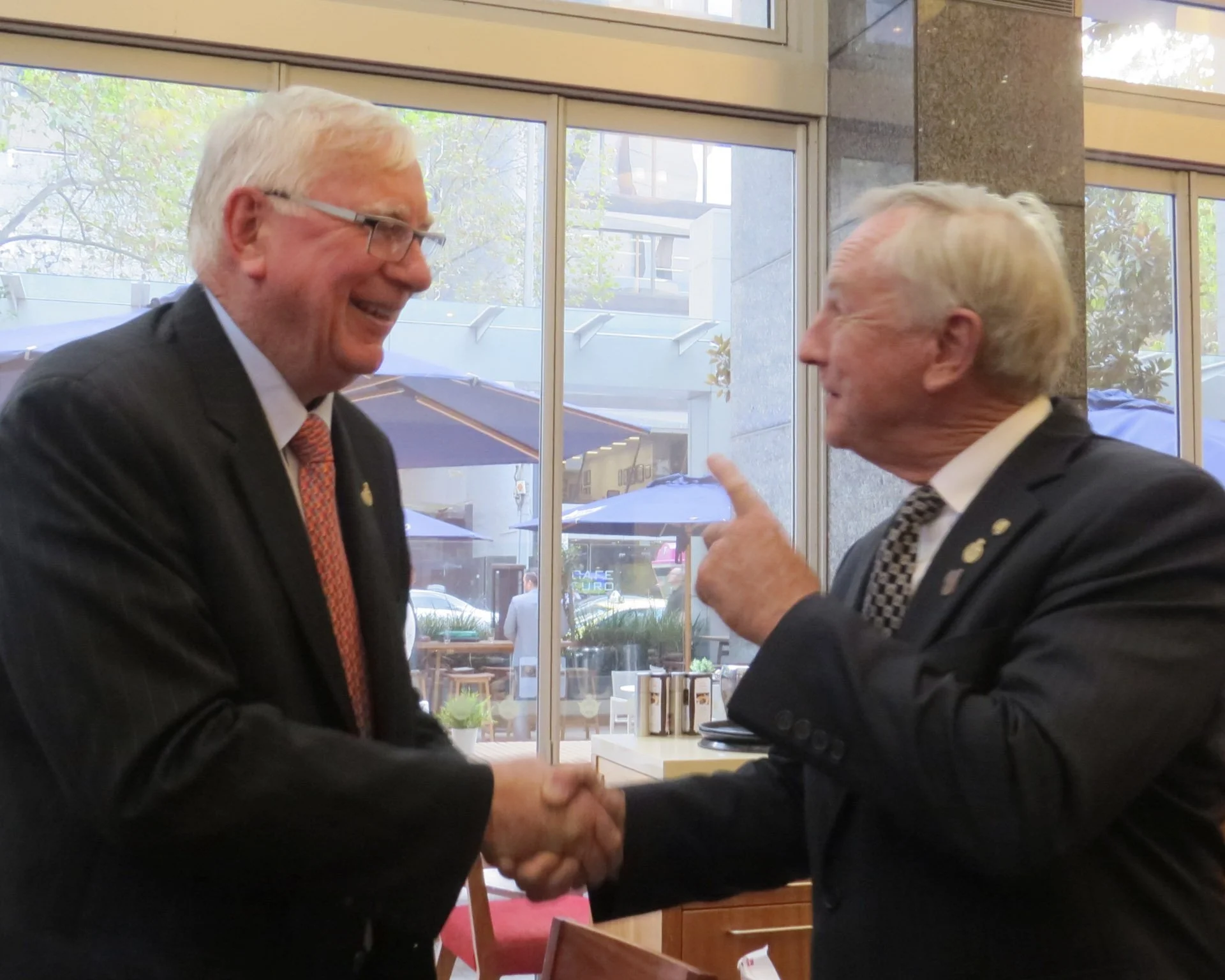 Two men in suits shaking hands and smiling inside a building with large windows, with outdoor seating visible through the windows.