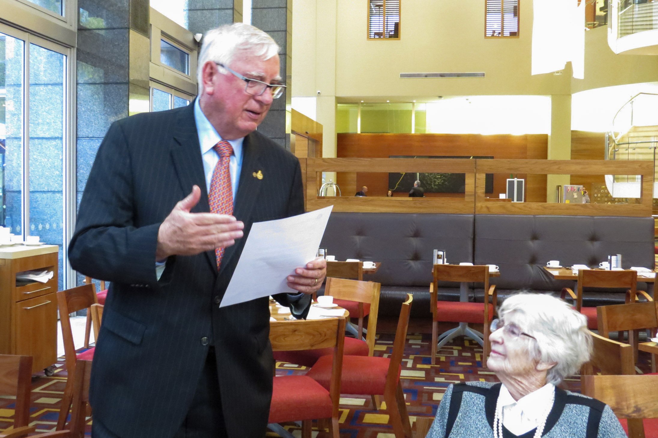 An elderly woman with white hair, glasses, and pearl necklace looks up at a man in a black suit holding a piece of paper, inside a restaurant with wooden chairs and tables.