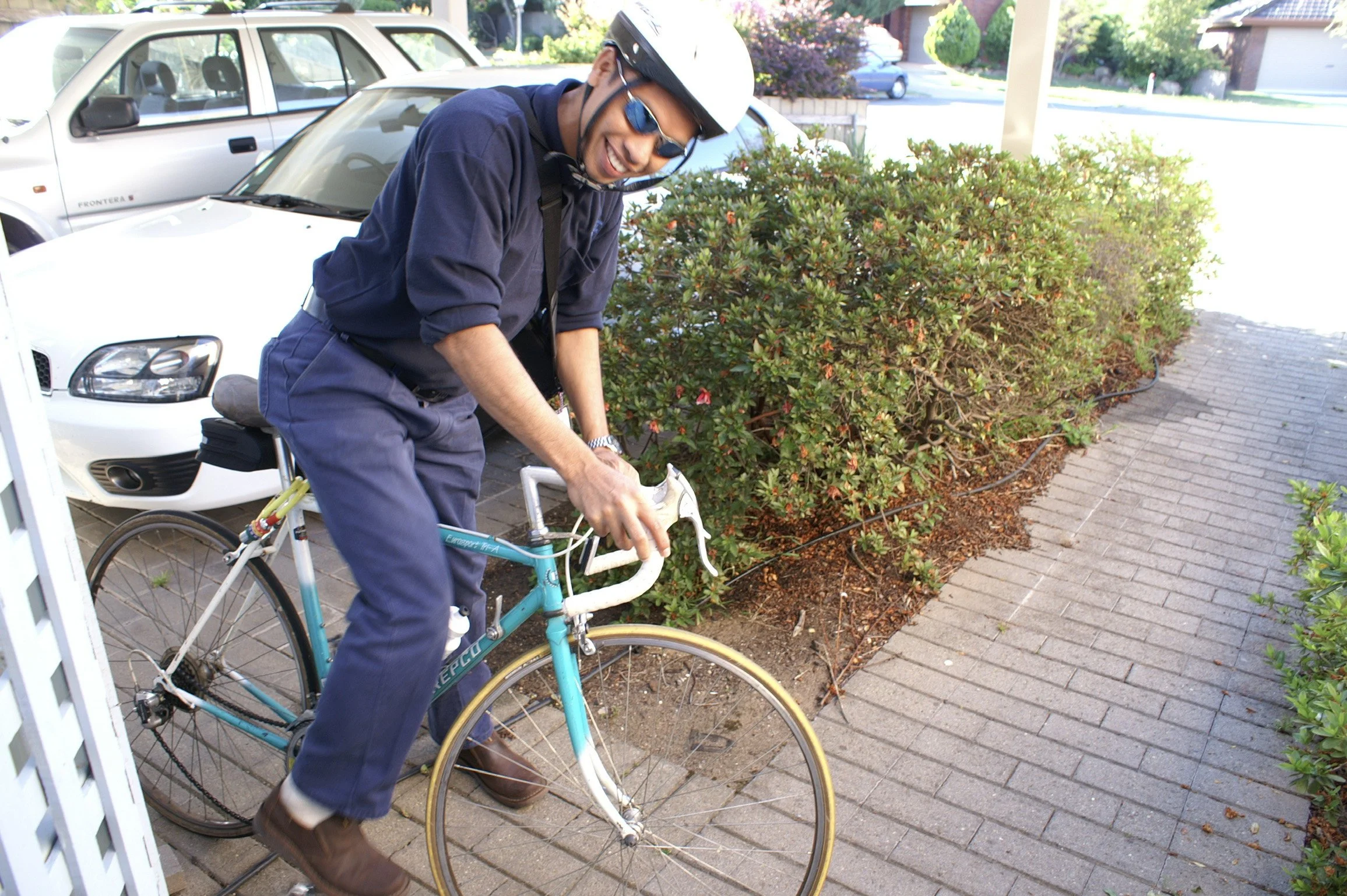 A man wearing a white helmet, sunglasses, and navy blue clothing rides a vintage teal and white bicycle on a brick sidewalk next to bushes and parked cars.