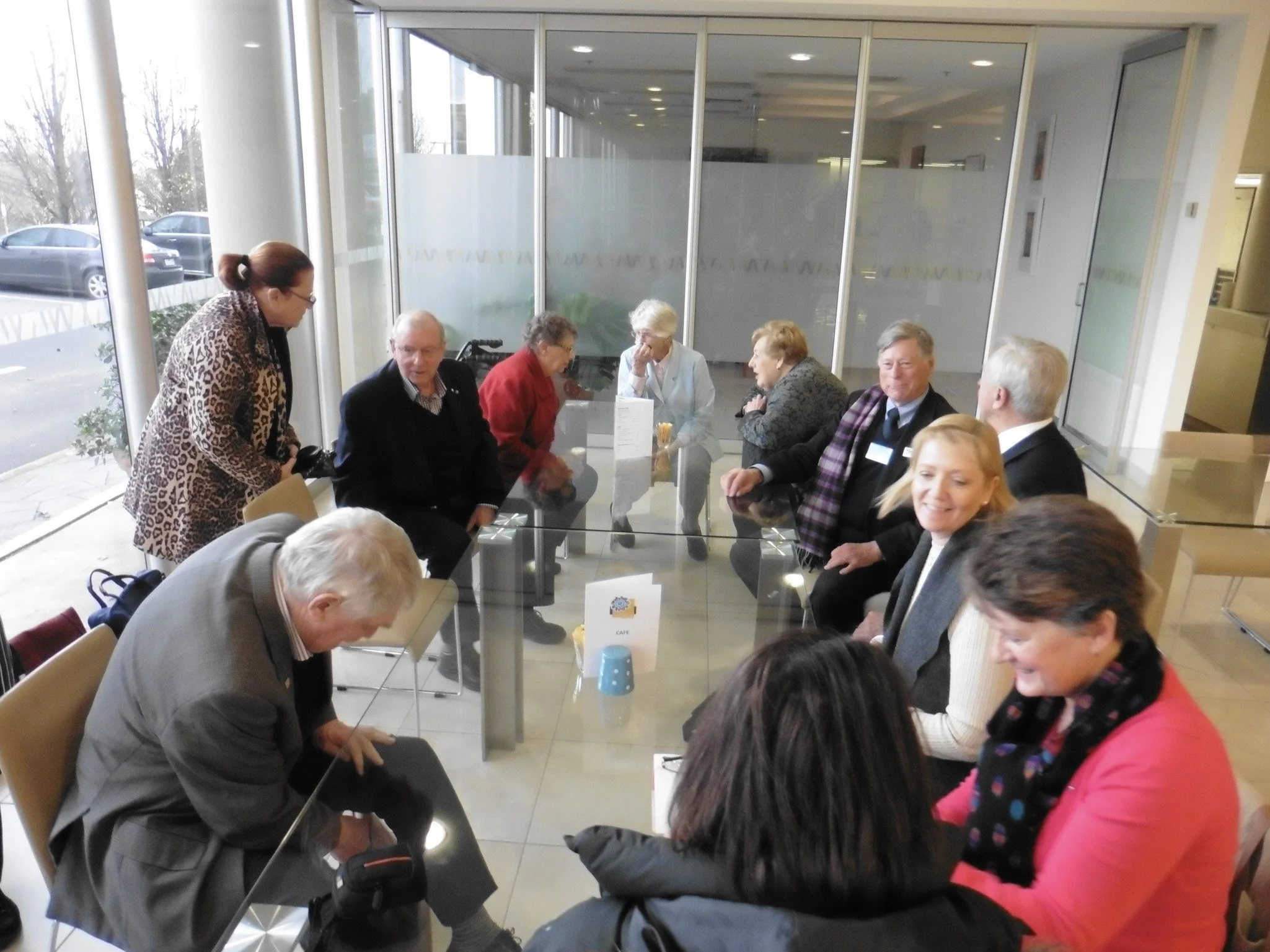 A group of elderly and middle-aged people sitting around a glass conference table in a modern office with large windows, during a meeting or social gathering.