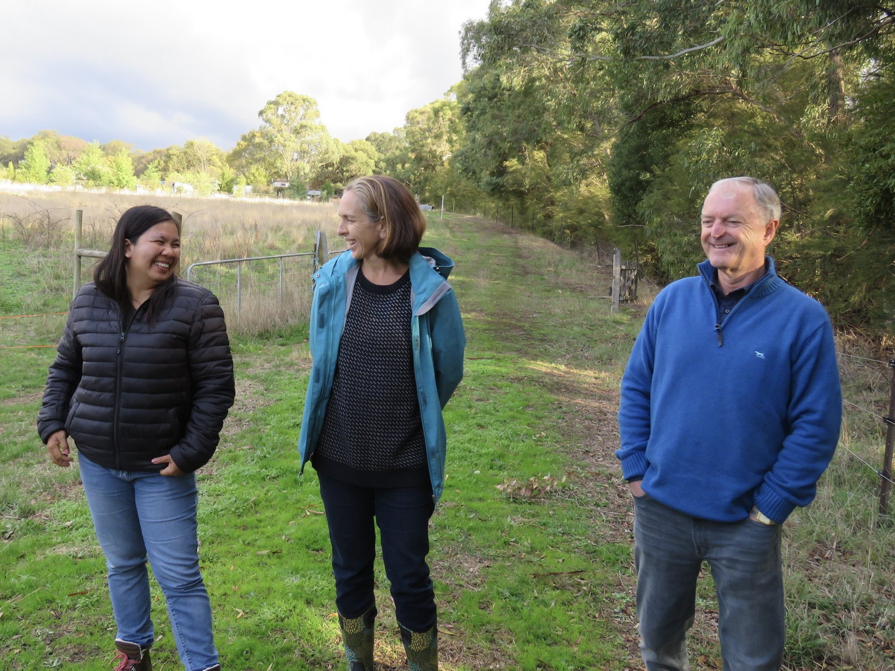 Three people standing on a grassy trail outdoors, smiling and laughing, with trees and a fenced-off field in the background.