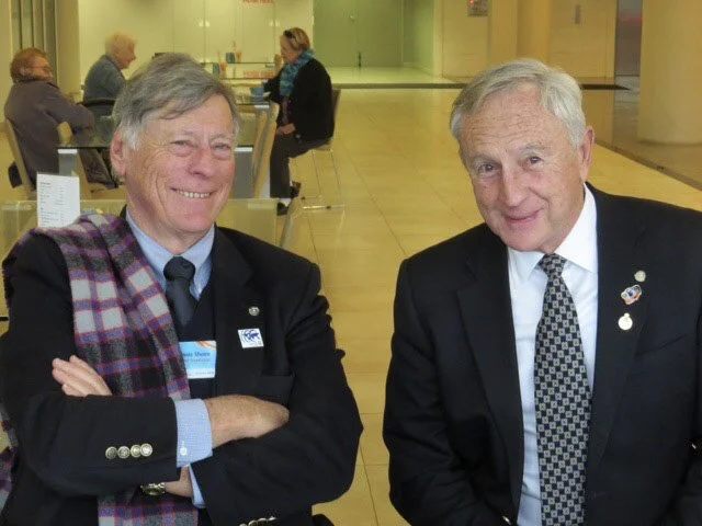 Two elderly men in suits smiling at the camera in an indoor setting, with several people visible in the background.