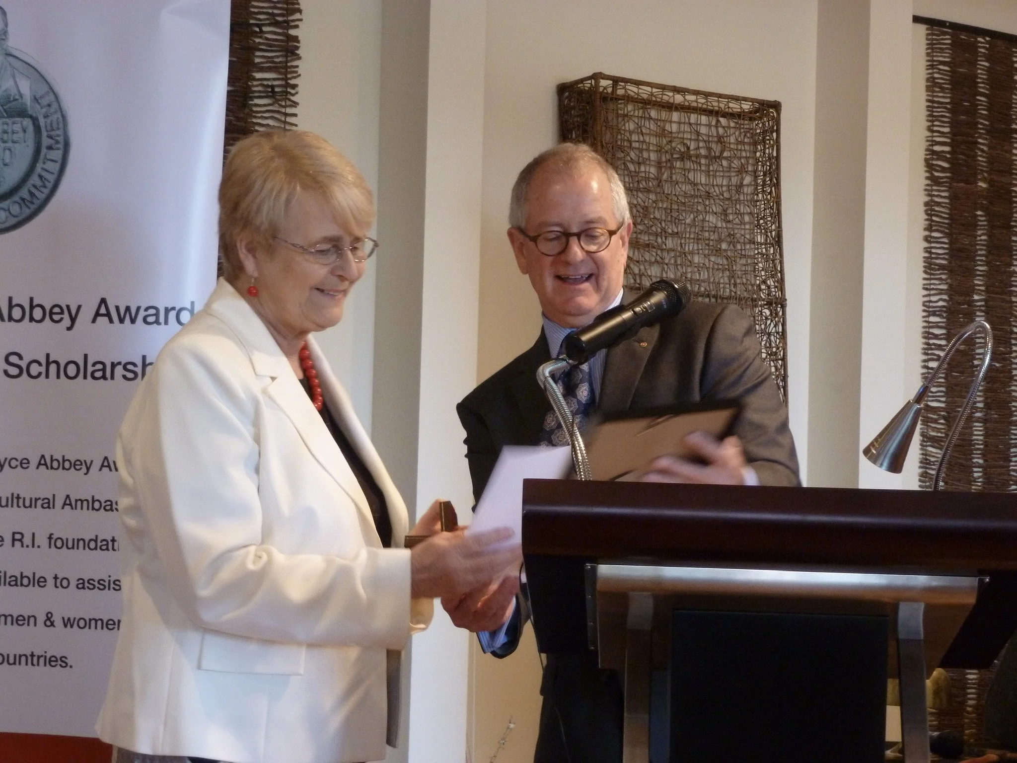 An elderly woman in a white blazer and elderly man in a suit at a podium, receiving an award or certificate with a microphone.