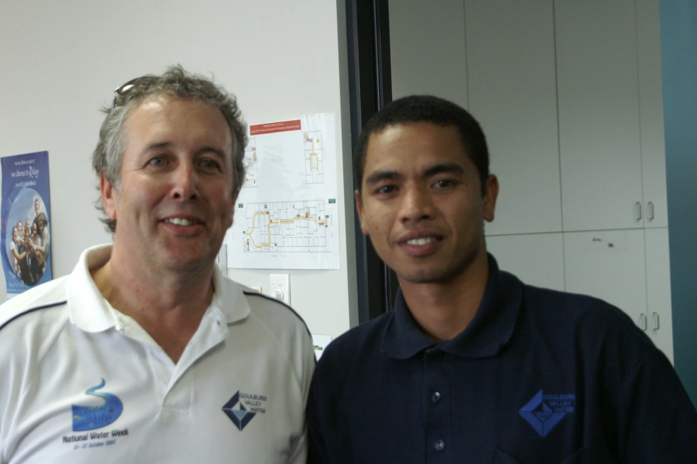 Two men posing for a photo indoors. The man on the left has gray, curly hair and is wearing a white polo shirt with logos related to water and a water organization. The man on the right has short black hair and is wearing a dark blue polo shirt with 