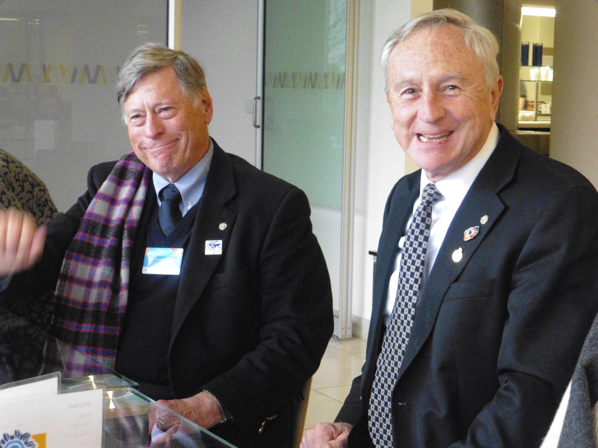 Two older men in suits sitting at a table, smiling for the camera. One is wearing a dark suit and tie with a lapel pin and the other a scarf, light blue shirt, and a badge. The background shows a glass door and an indoor setting.