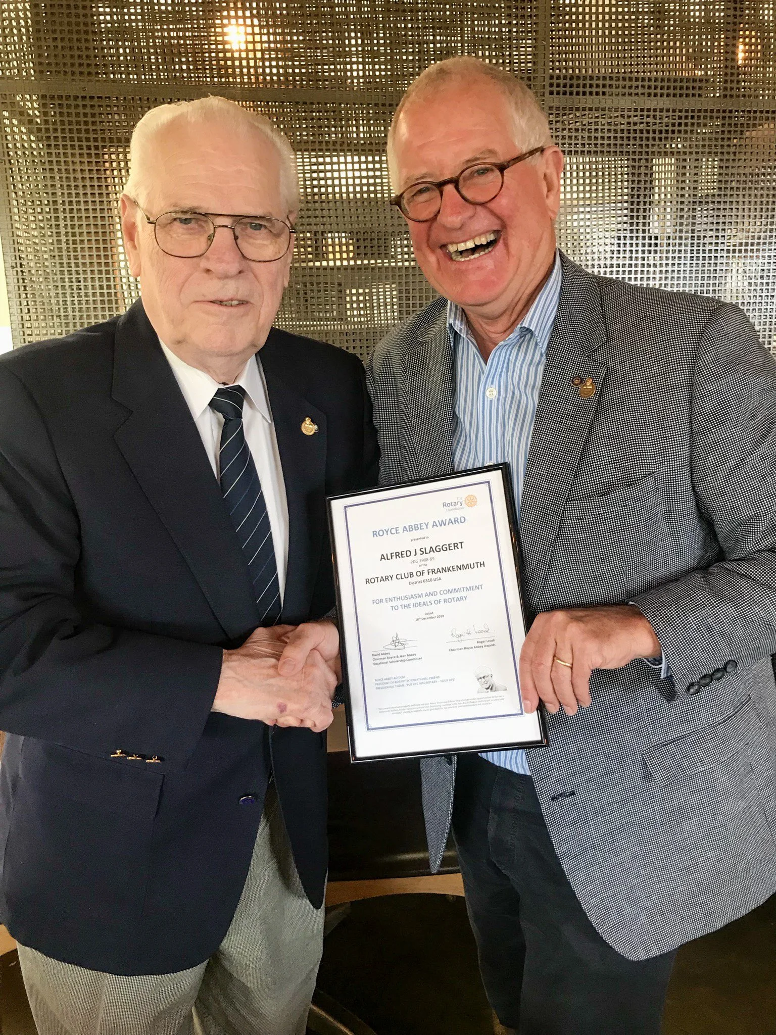 Two men in suits shaking hands and holding a framed award certificate at a recognition event.