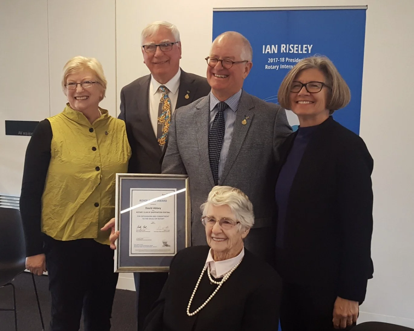 Group of five people, four standing and one seated, at a recognition ceremony, with one holding a framed certificate and a blue banner in the background reading 'Ian Riseley, 2017-18 President, Rotary International.'