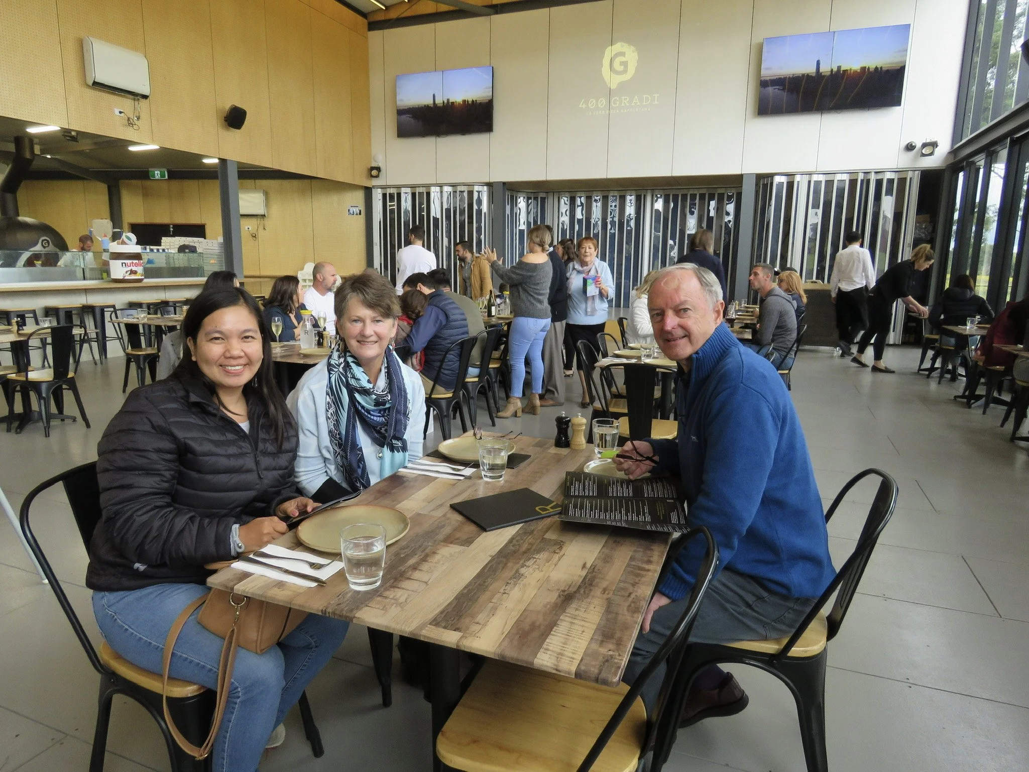 Three people sitting at a wooden table in a busy modern restaurant or cafe, smiling at the camera. Two women and one man, with glasses of water and menus on the table. Other diners and staff are visible in the background.