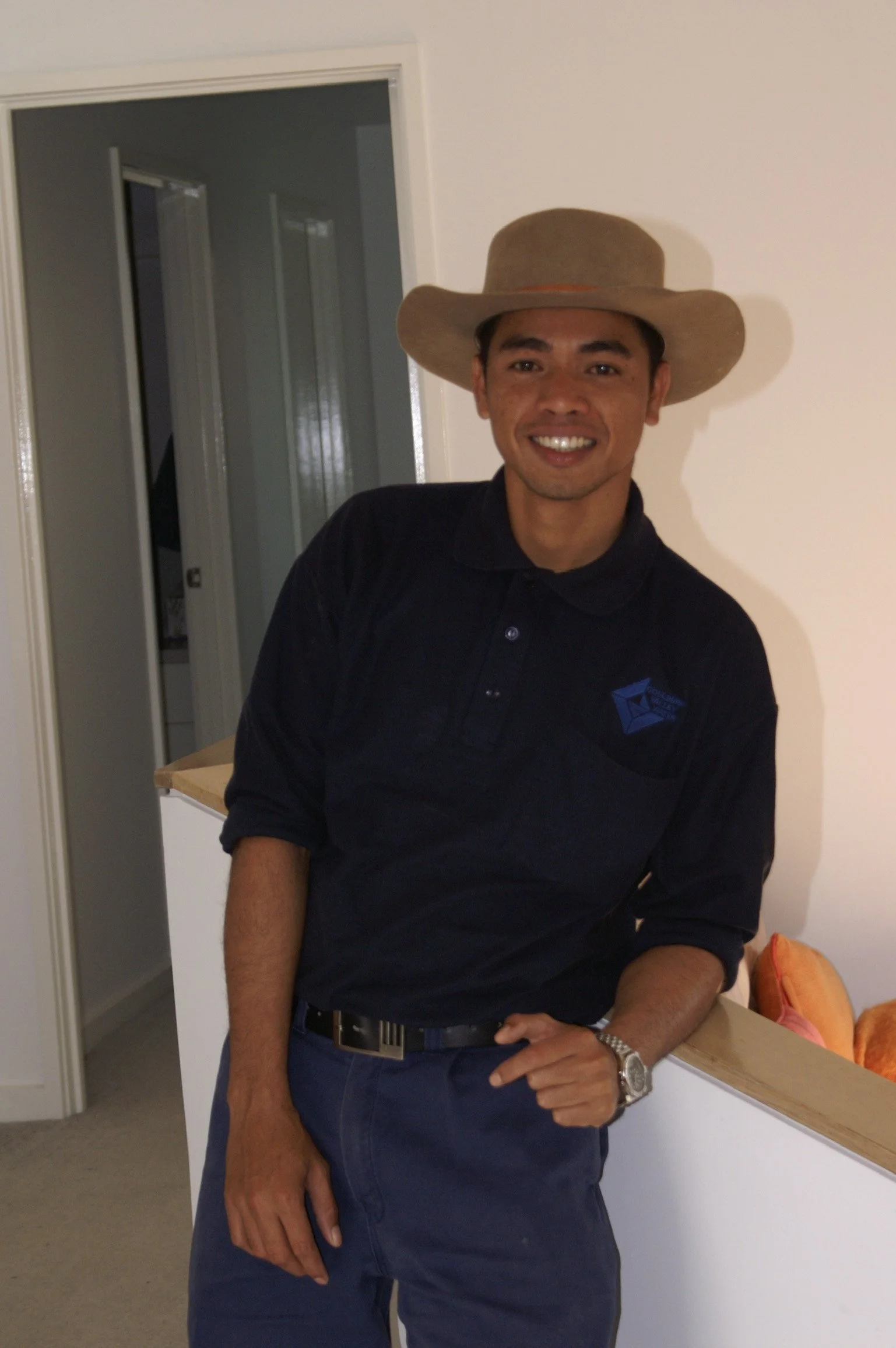 A man smiling, wearing a wide-brimmed hat, a black polo shirt with a logo, and a wristwatch, standing in a room with a beige wall and an open door in the background.