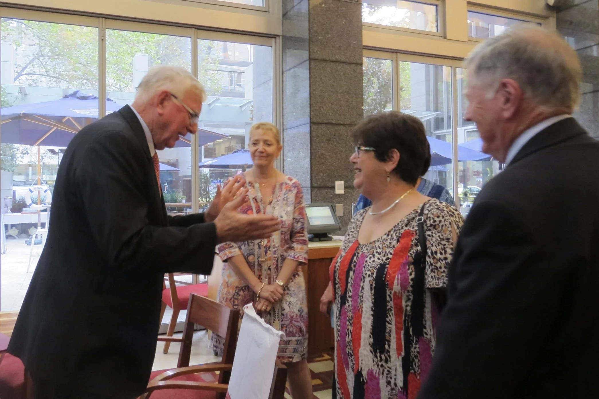 Four people, two men and two women, are standing and engaging in conversation inside a building with large glass windows and doors, lighting, and a view of umbrellas and outdoor seating outside.