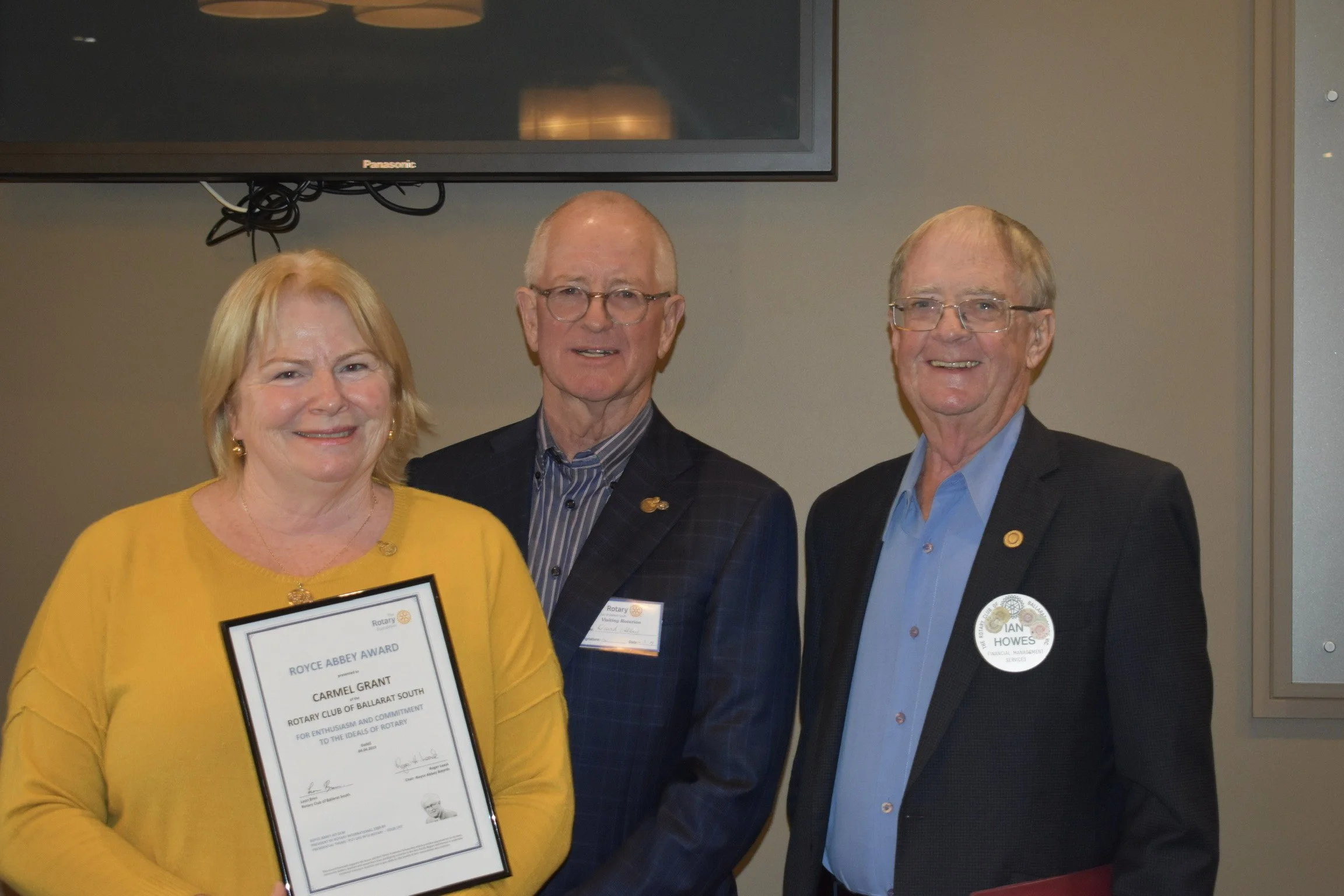 Three people at a formal event, one woman holding a framed award certificate, smiling for the photo, two men standing beside her also smiling, with a large flat-screen TV on the wall behind them.