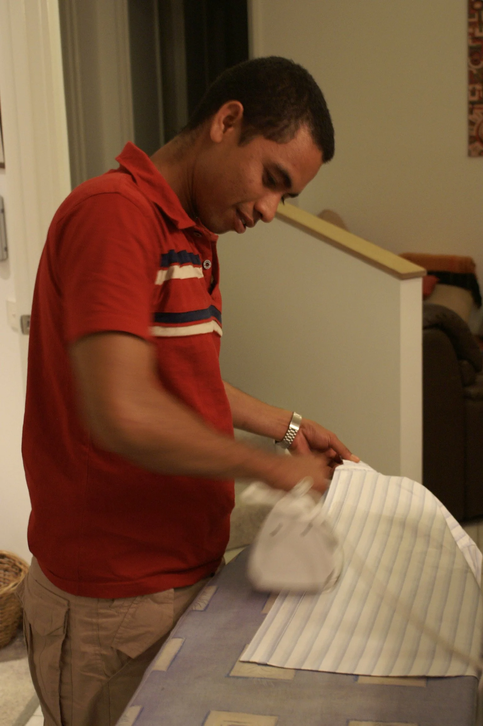 A young man in a red polo shirt and beige pants is ironing a striped shirt on an ironing board in a home setting.