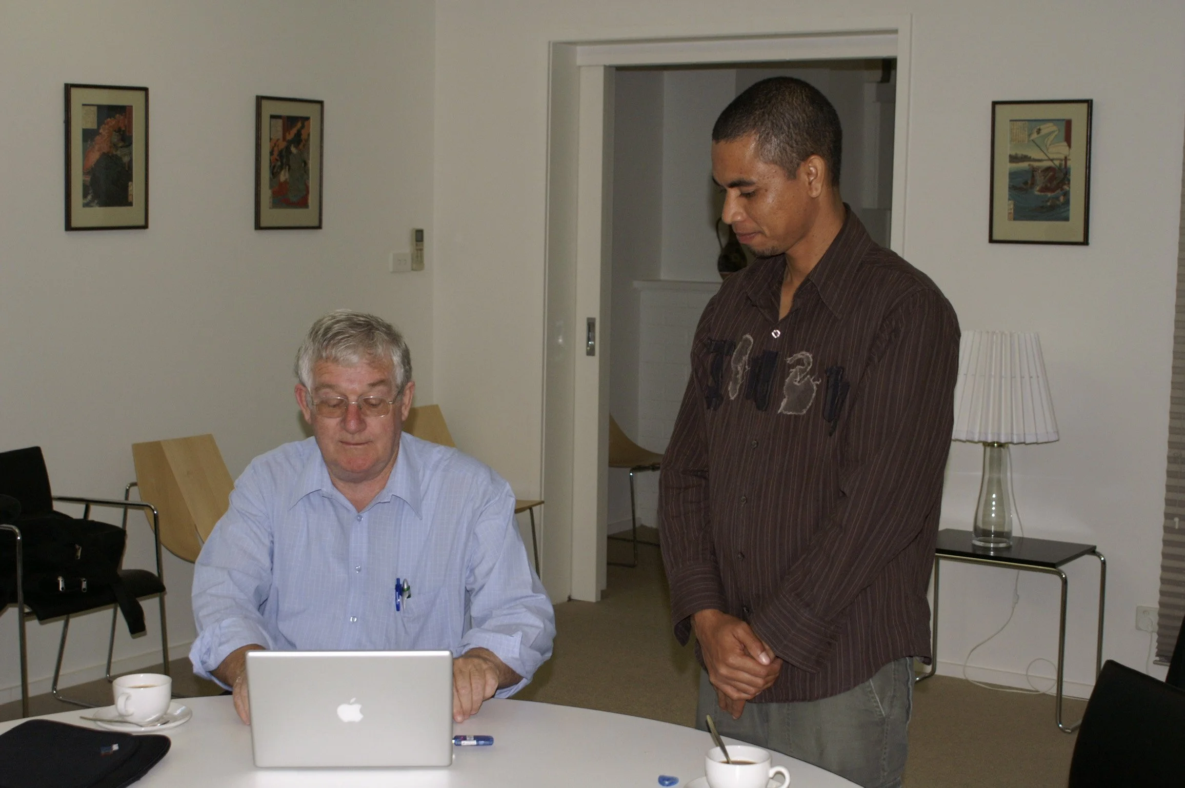Two men in an office or conference room have a discussion. One man with gray hair and glasses is seated, looking at a laptop, and the other man with short dark hair is standing, looking down.