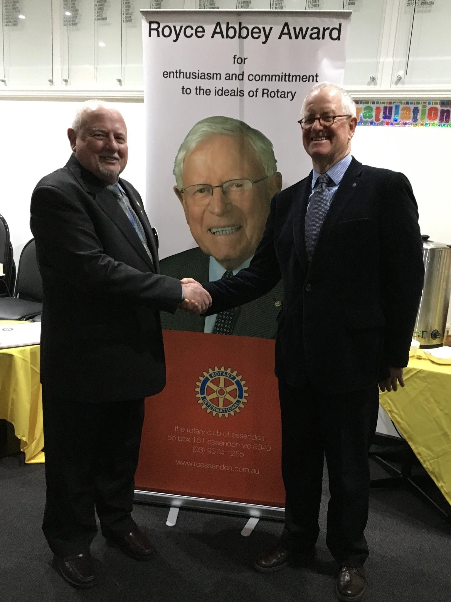 Two men in suits shaking hands at an awards ceremony with a large portrait of a smiling older man in the background and a banner that reads 'Royce Abbey Award for enthusiasm and commitment to the ideals of Rotary'.
