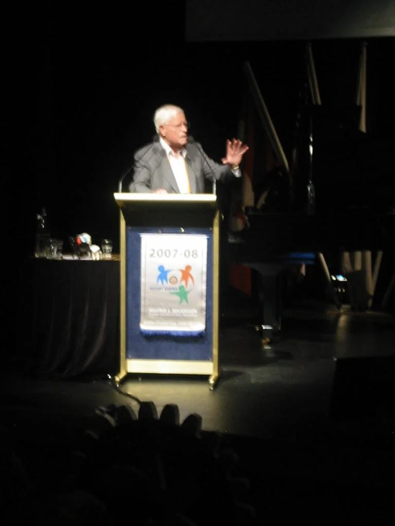 A man in a gray suit speaking at a podium on a dark stage, with a poster on the front of the podium indicating the years 2007-08 and featuring colorful human figures.