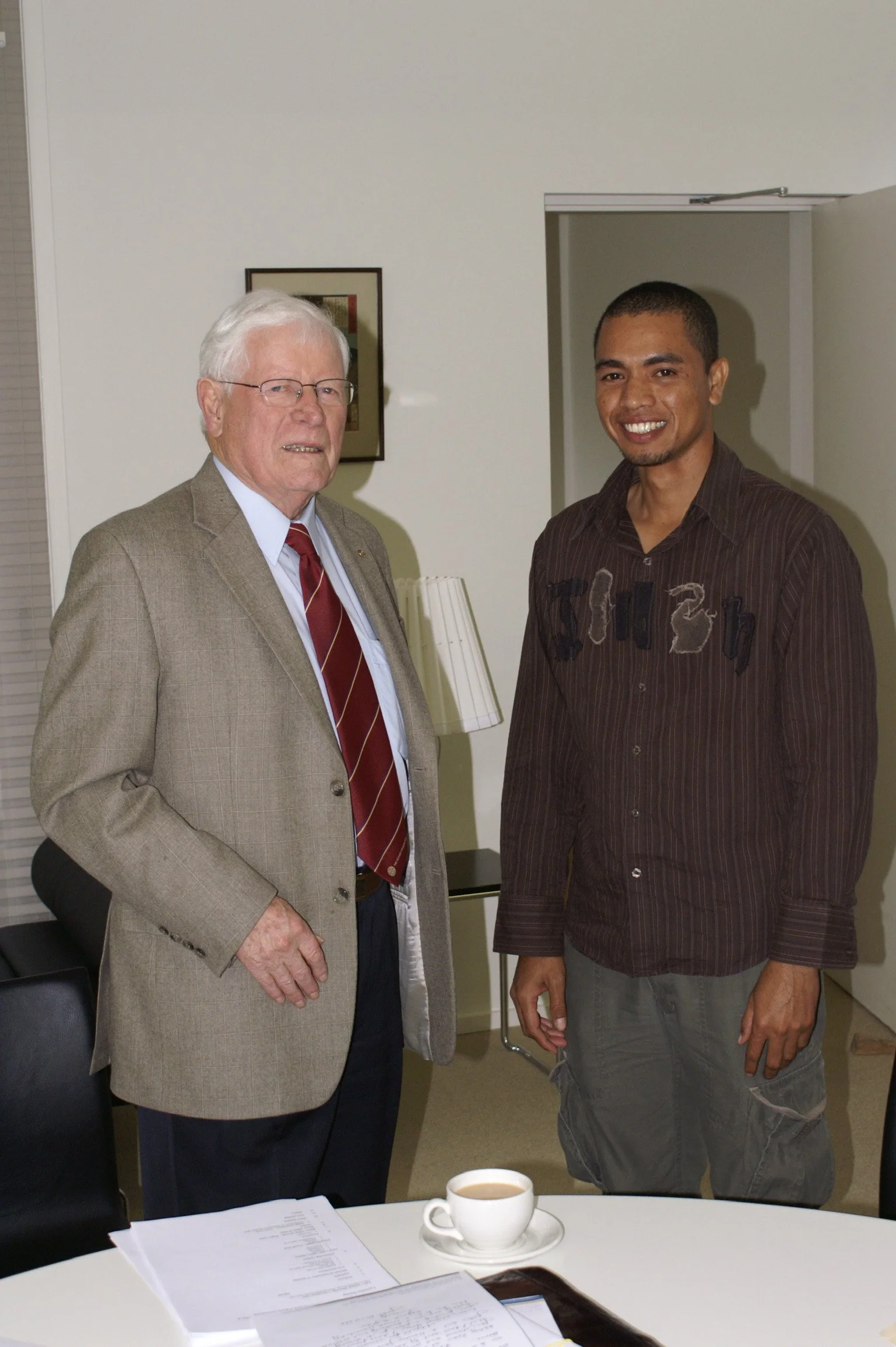Two men standing side by side indoors. One is elderly with white hair, glasses, wearing a beige blazer, shirt, and striped tie. The other is younger with short black hair, wearing a striped brown shirt and cargo shorts. There is a table with a cup of