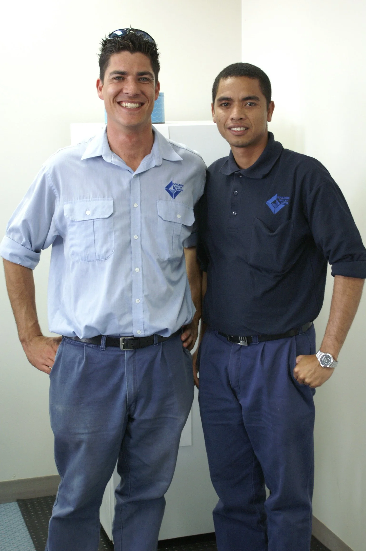 Two men standing side by side indoors, smiling at the camera, wearing company uniforms in light blue and navy blue with logos.