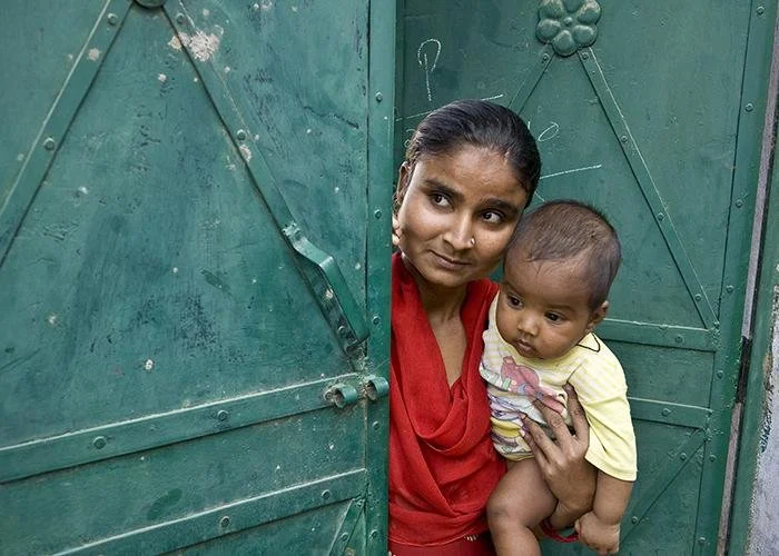 A woman with dark hair and a red shawl holds a young child with a yellow shirt, peeking out from behind a green metal door.
