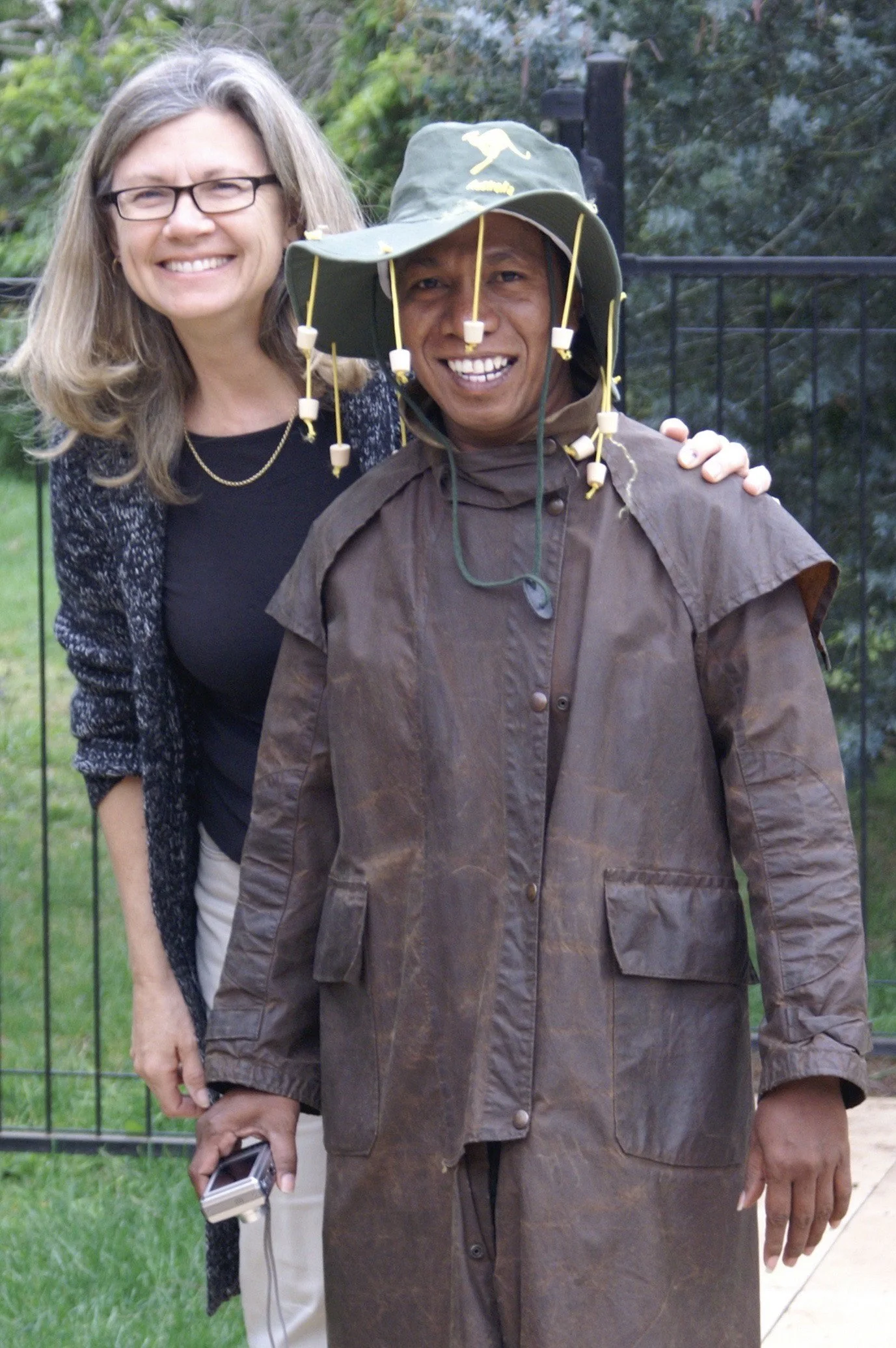 A woman and a man smiling together outdoors, with the woman resting her hand on the man's shoulder. The man is wearing a wide-brimmed hat with hanging cords and a brown, weathered jacket, while the woman has long gray hair, glasses, and a black top w