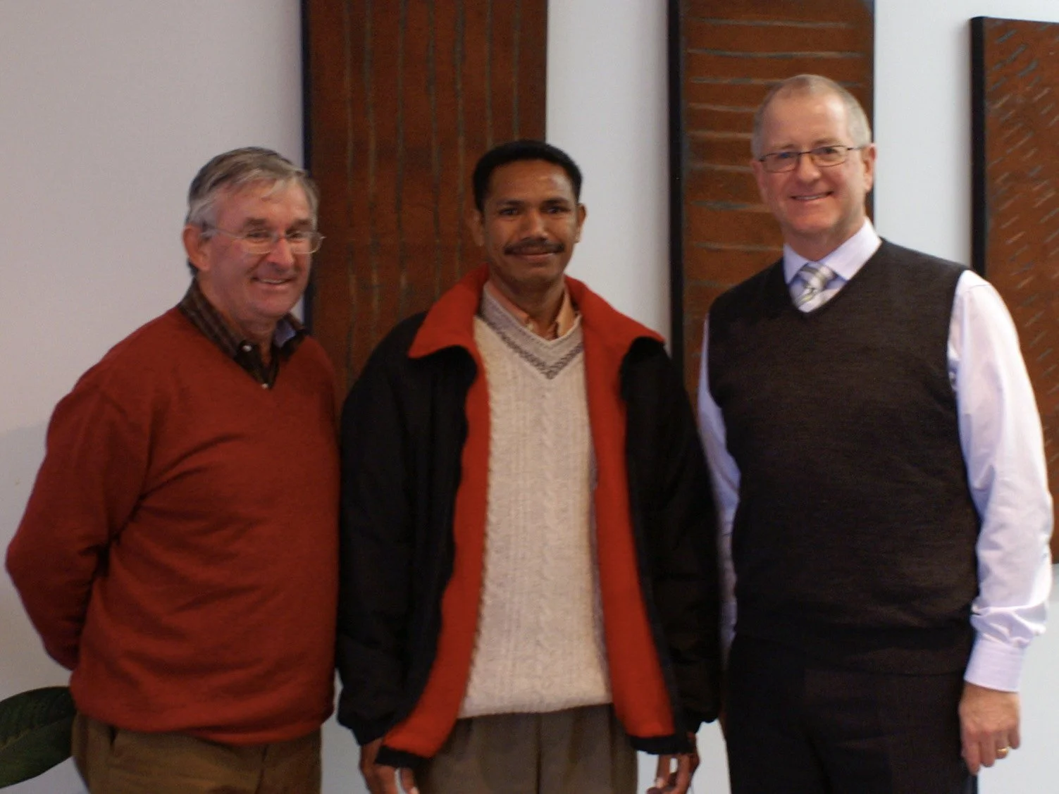 Three men standing together indoors, smiling, with a brick wall and white pillar in the background.