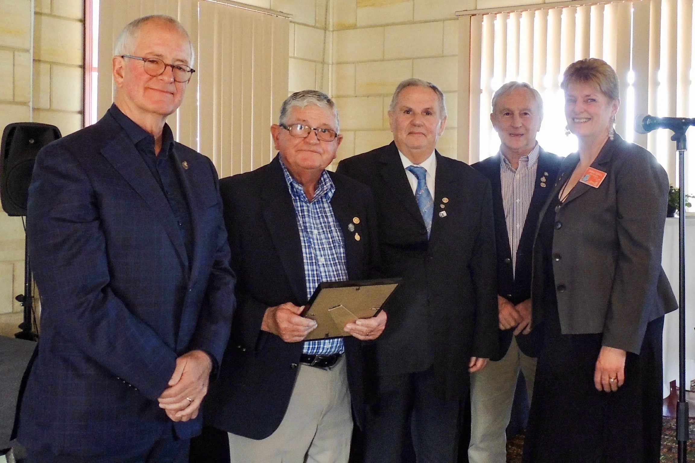 Six individuals in formal attire standing indoors, with one holding a framed certificate or plaque, all smiling for the photo.