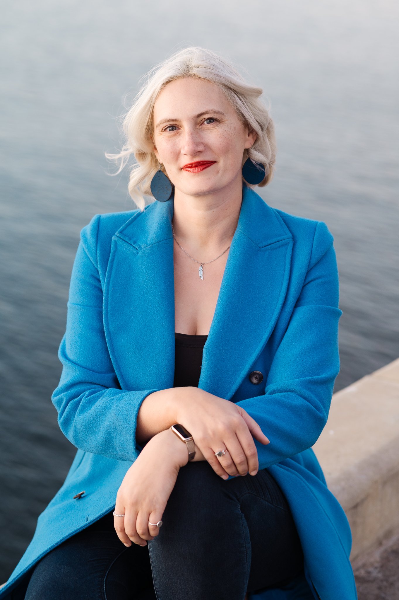 A woman with platinum blonde hair wearing a vibrant blue blazer and matching earrings, sitting by a body of water with a slight smile.