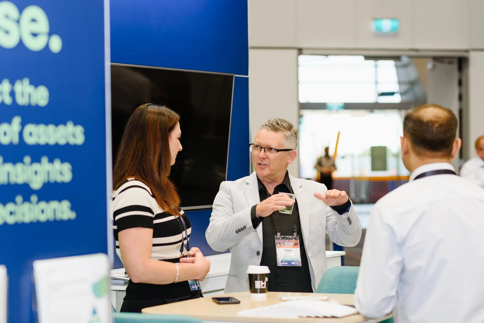 A man in a light gray blazer and glasses is speaking to a woman with long brown hair, wearing a black and white striped shirt, at a conference. Another man in a white shirt with a black lanyard is also part of the conversation. They are standing around a table with a coffee cup and phone.